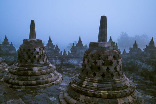 Ancient stone stupas at Borobudur Temple emerge from the morning mist in Central Java, Indonesia. The atmospheric scene showcases one of the world's most renowned Buddhist monuments