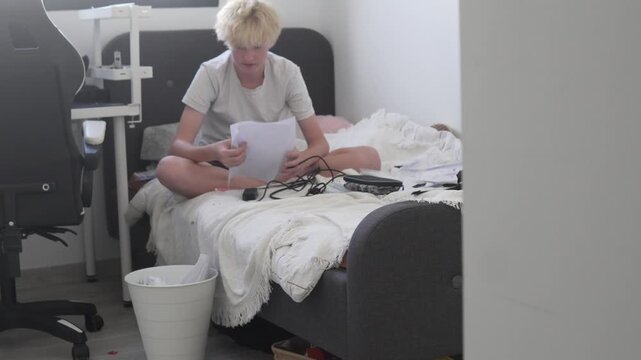 Blond boy sorting school papers at home after the end of the school year and throwing unnecessary documents into a trash bin. Teenager organizing his room, cleaning up study materials and decluttering