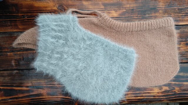 Horizontal flat lay of a fluffy light grey angora knitted hood over a beige wool hood. Handmade winter balaclavas on a dark rustic wooden surface. Cozy accessories