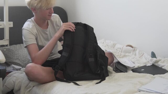Teenage boy sitting on a bed at home and organizing his school backpack. Student unpacking school supplies and papers in a bedroom after classes, preparing, cleaning, and sorting study materials