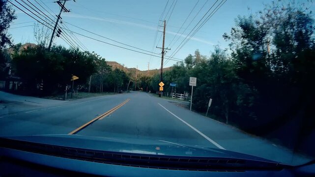 Dashcam view driving Mulholland Highway. Winding road, double yellow line, green hills, dense trees. Utility poles, power lines roadside. Distant mountain range under clear blue sky. Scenic journey th