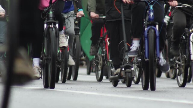 Large gathering of individuals participating in a bicycle parade, riding various bikes and one electric scooter on a city road. Focus on motion blur of wheels and legs, creating sense of speed and act