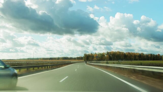 POV shot of country highway curving through scenic autumn landscape. Golden trees line road under bright blue sky with fluffy white clouds. Guardrails visible on side of highway. Open road travel jour