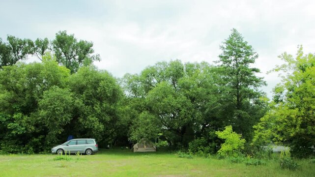 Full shot of a silver minivan parked beside a green camping tent at border of lush forest, open grassy field leading to trees, cloudy day, summer nature exploration.