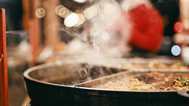 Street Food Cooking On A Big Skillet With Steam Against The Evening Lights Of The Fair