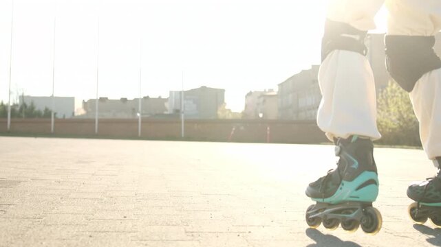 A girl wears white pants and black and teal roller skates. She skates outdoors on light pavers in bright sun.