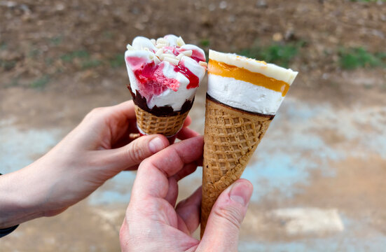 Two ice cream cones held by hands with different flavors in a natural setting during daylight hours