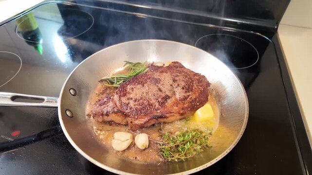 A professional chef performs butter basting on a searing ribeye steak in a stainless steel pan, incorporating aromatic garlic, rosemary, and thyme to enhance flavor and finish the cooking process.