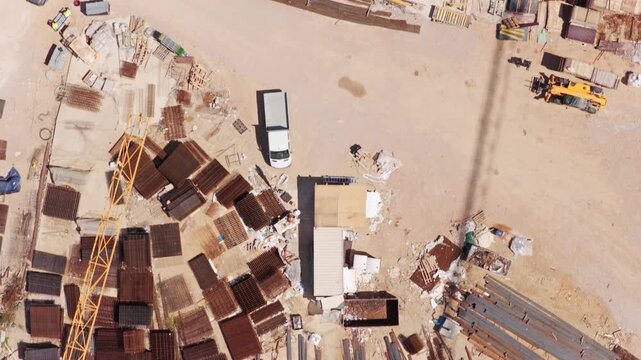 Top-down aerial view of a construction site storage area with organized piles of rusty rebar, steel beams, and various building materials scattered on the sandy ground in bright sunlight