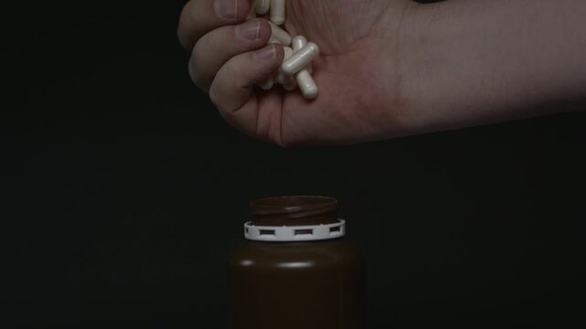 Close-up of a hand filling a brown medicine bottle with capsules in slow motion. Some pills fall outside the container on a black background.