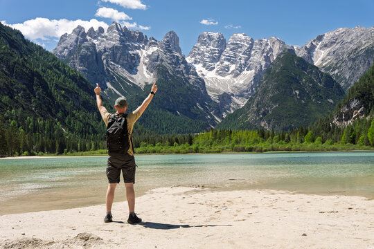 Male tourist with backpack standing with raised arms in celebration on the sandy shore of Lake Landro facing majestic rocky mountain peaks, success and solo wilderness exploration concept