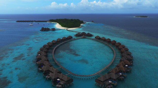 Aerial pan and descent show Maldives ring shaped overwater villas, plunge pool terraces, wooden boardwalks, long jetty, shallow turquoise lagoon, reef, boats, islet.