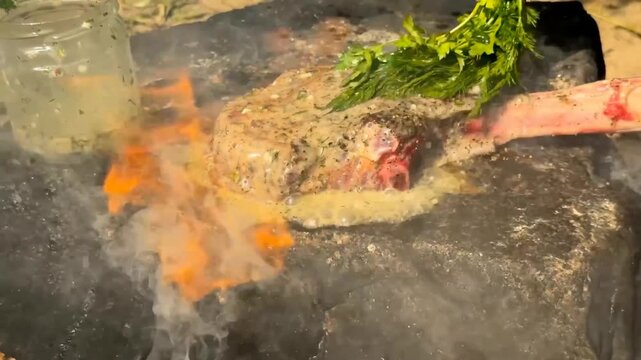 A person uses a bundle of fresh herbs to continuously baste a sizzling tomahawk steak with a rich butter sauce on a heated stone slab over an open wood fire during an outdoor cooking session