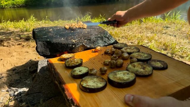 A person transfers grilled mushrooms, eggplant slices, and a large tomahawk steak from a hot stone onto a rustic wooden serving board in a natural riverside setting during the bright daylight hours
