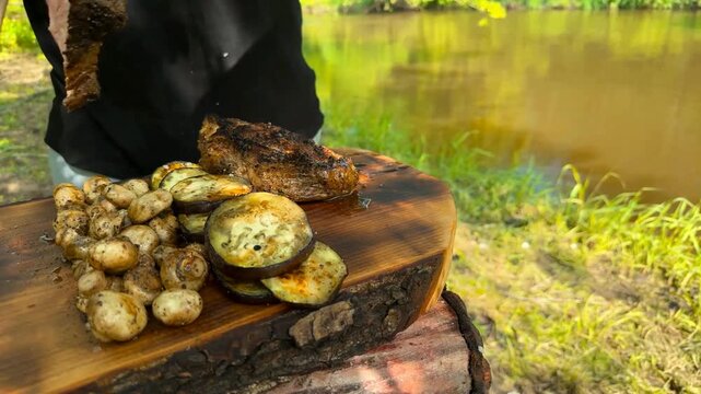 A person carves a cooked tomahawk steak on a wooden board next to grilled mushrooms and eggplant slices in a natural riverside setting during the bright daylight hours