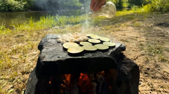 A person seasons mushrooms and eggplant slices with oil and spices while grilling them on a large flat stone over a campfire in a serene riverside environment during the bright daylight hours
