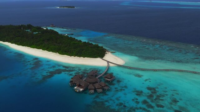 Aerial view of a Maldives resort island with circular water villas, long jetties, white sand, turquoise lagoon, coral patches, and a slow pan in bright daylight.