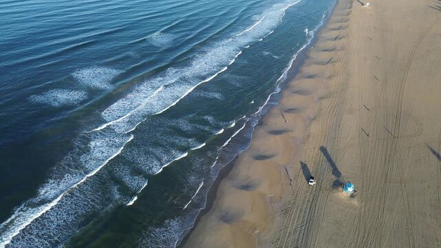Lifeguard stations long tire tracks trace wide Huntington Beach sands, framing transition between deep surf dry dunes. Soft light casts lengthy shadows across this calm, expansive region in CA