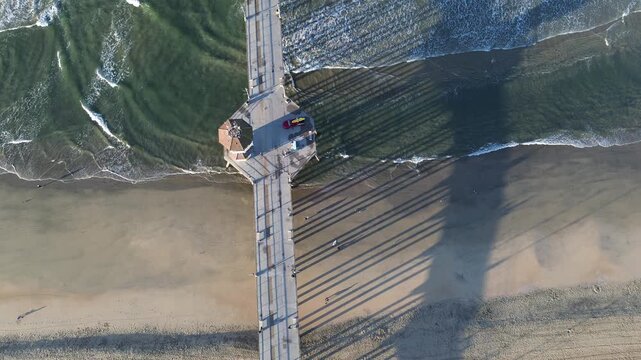Direct overhead views capture geometric precision of Huntington Beach Pier bisects shore. Long, dark structural shadows stretch across sand toward rhythmic, incoming ocean surf, Orange County, CA