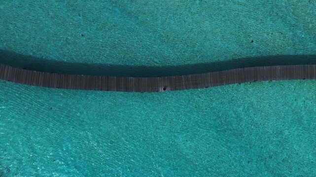 Aerial top down view shows a sinuous wooden boardwalk in the Maldives, shallow turquoise lagoon, rippling sand, coral patches, and a lone figure under bright daylight.
