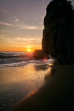 Stimmungsvoller Sonnenuntergang an einem Sandstrand auf Korfu mit gro&szlig;en Felsen, sanfter Brandung und Reflexionen im nassen Sand.