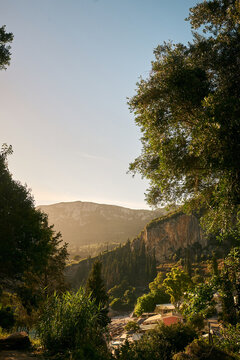 Malerischer Blick durch &uuml;ppige gr&uuml;ne B&auml;ume auf eine Berglandschaft, steile Klippen und ein kleines K&uuml;stendorf auf Korfu im goldenen Licht