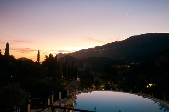 Stimmungsvoller Sonnenuntergang &uuml;ber einer Berglandschaft auf Korfu, gesehen &uuml;ber einen beleuchteten Swimmingpool mit spiegelndem Wasser.