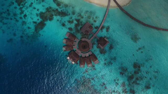 Aerial top down view of a Maldives resort shows circular overwater villas, a curved jetty to a white sand island, turquoise lagoon, coral patches, and plunge pools in daylight.