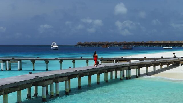 Aerial Maldivian resort shows a curving wooden jetty to a white beach. A woman in a red dress walks as yachts and boats bob in deep water under midday light.