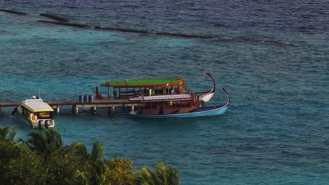 Aerial view shows two dhoni boats, blue and white hulls, tied to a wooden jetty in the Maldives. Crew move on deck, reef booms trace the outer reef in warm light.