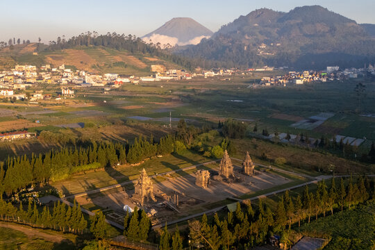 Aerial view of the ancient Arjuna Temple complex surrounded by lush trees and agricultural fields with mountains in the background in Dieng Kulon, Jawa Tengah, Indonesia.