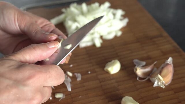 Close-up  a woman&rsquo;s hands peeling garlic with a knife. The woman is cooking in her kitchen.