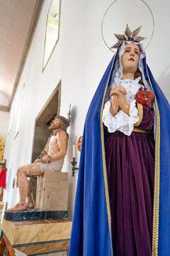 Our Lady of Sorrows with her back to Jesus Christ seated in suffering. The statue presents typical characteristics of Marian iconography, including a blue mantle and a visible ex-voto heart.
