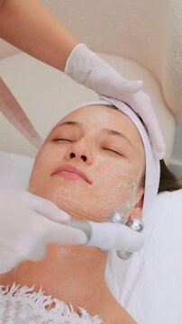 Young woman relaxing during a professional facial treatment at a beauty salon, where a beautician uses a specialized roller massager for lymphatic drainage and skin lifting on her face. Vertical video