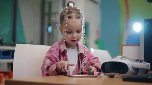 Concept technology in education school, young girl tapping tablet and reaching for vr headset on table in colorful indoor playroom with smooth lateral pan.