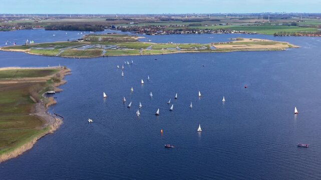 Aerial view of a sailboat regatta on a wide lake in spring. Drone landscape of numerous sailing yachts, water sports, green polder islands, and coastal villages in the Netherlands, panorama.
