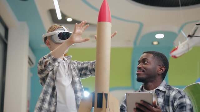 Boy interacts with vr headset and cardboard rocket in space classroom makerspace while mentor observes and records on tablet, technology in education of school.