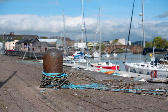 A rusty bollard wrapped with blue mooring rope sits on stone quay while sailboats and yachts rest in the busy harbor, a nautical atmosphere, seaside leisure and maritime infrastructure