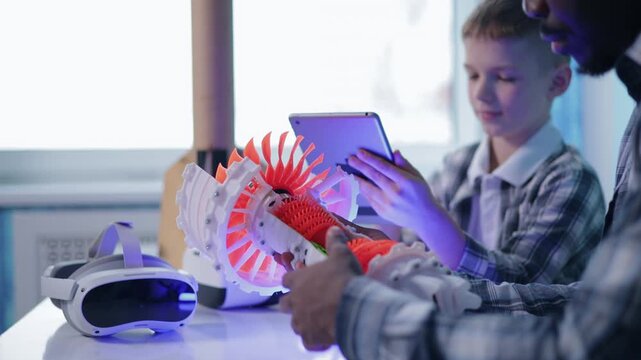 Boy and adult examining 3d printed turbine model on classroom table with tablet interaction, concept technology in education school.