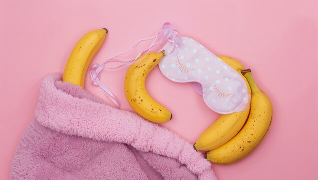Pink sleep mask with eyelashes placed on a soft pink blanket alongside several ripe bananas arranged artistically on a pastel pink background