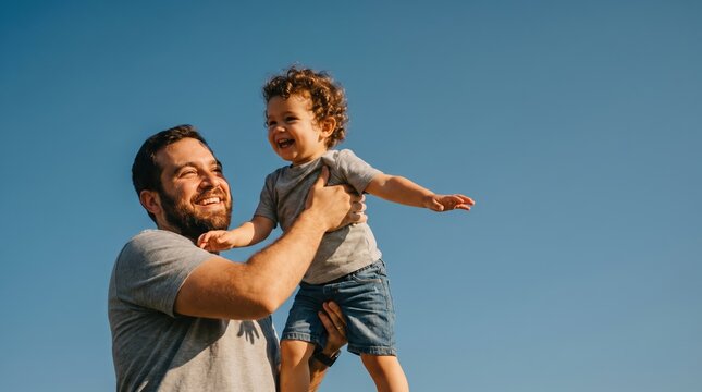 Father lifting laughing toddler son in the air against clear blue sky outdoors for family bonding and parenting lifestyle content
