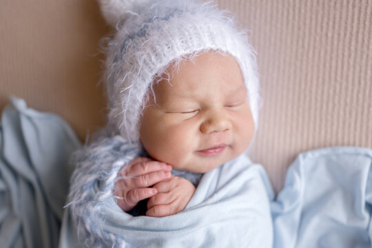 Sweet newborn baby sleeps soundly. Wrapped in soft blue swaddle. Knit hat adorns head. Professional newborn photoshoot.
