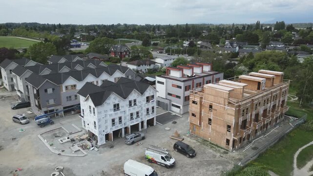 High-angle aerial perspective pulls back across multi-phase residential development project showcasing new townhouses ranging from raw wooden framing to completed white siding black shingled roofs