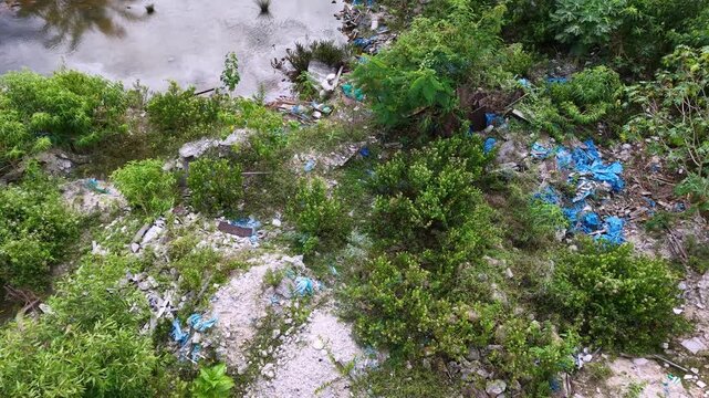 Drone footage shows a riverside outdoor rubbish dump scattered among vegetation and soil.