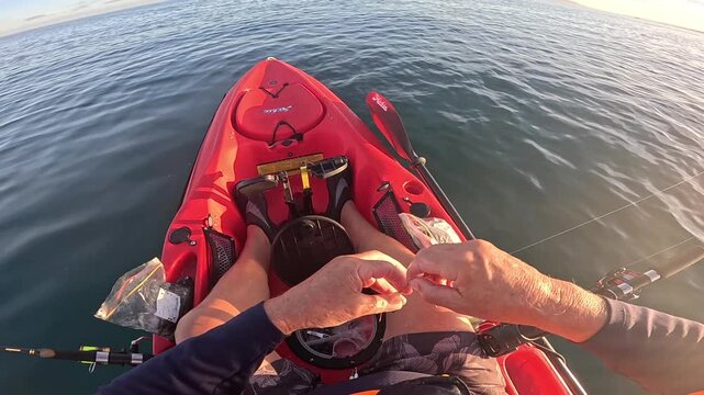 Angler sits in a red fishing kayak calmly changing lures over gentle evening sea water, viewed from first person cockpit angle.