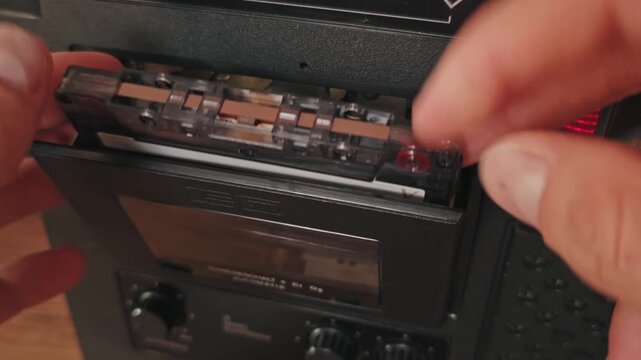 A hand inserts a transparent cassette into a vintage player deck and closes the mechanical door. The clear shell with visible internal reels highlights the industrial design of the audio device.