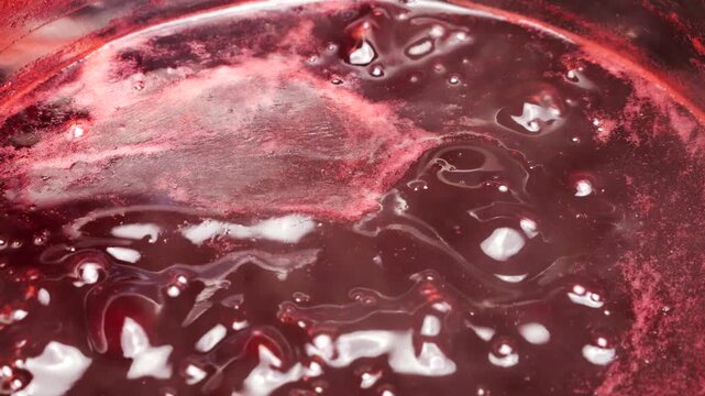 Boiling red currant jam bubbles in a saucepan during the home preparation process. Fresh berry confit cooks on the stove. Thick red jelly with small bubbles is simmering into sweet preserve.