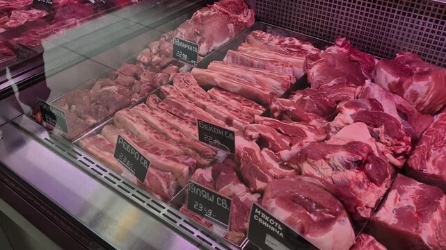 Various raw pork cuts like ribs and loin are neatly arranged in a refrigerated display case. The black price tags with Cyrillic text and pink lighting create a typical butcher shop atmosphere.