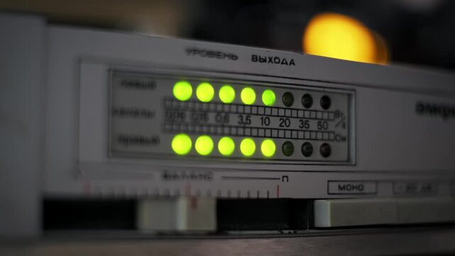 Close-up of a vintage analog amplifier output level meter with green LED lights fluctuating to music rhythm. The display features Russian text labels, creating a retro audio technology concept