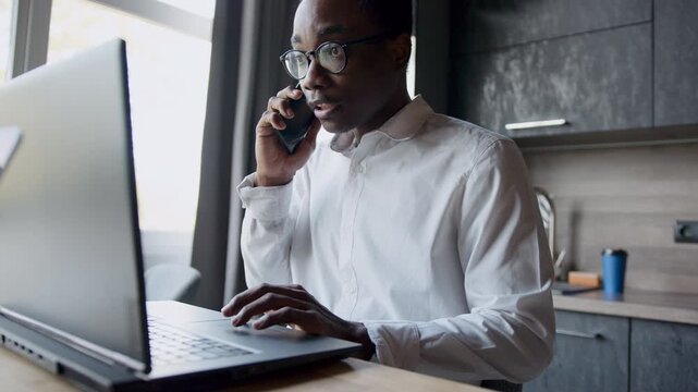 A professional Black man with glasses works intently on his laptop, engaged in a phone conversation from a stylish home office. He types while speaking, managing tasks efficiently during the day